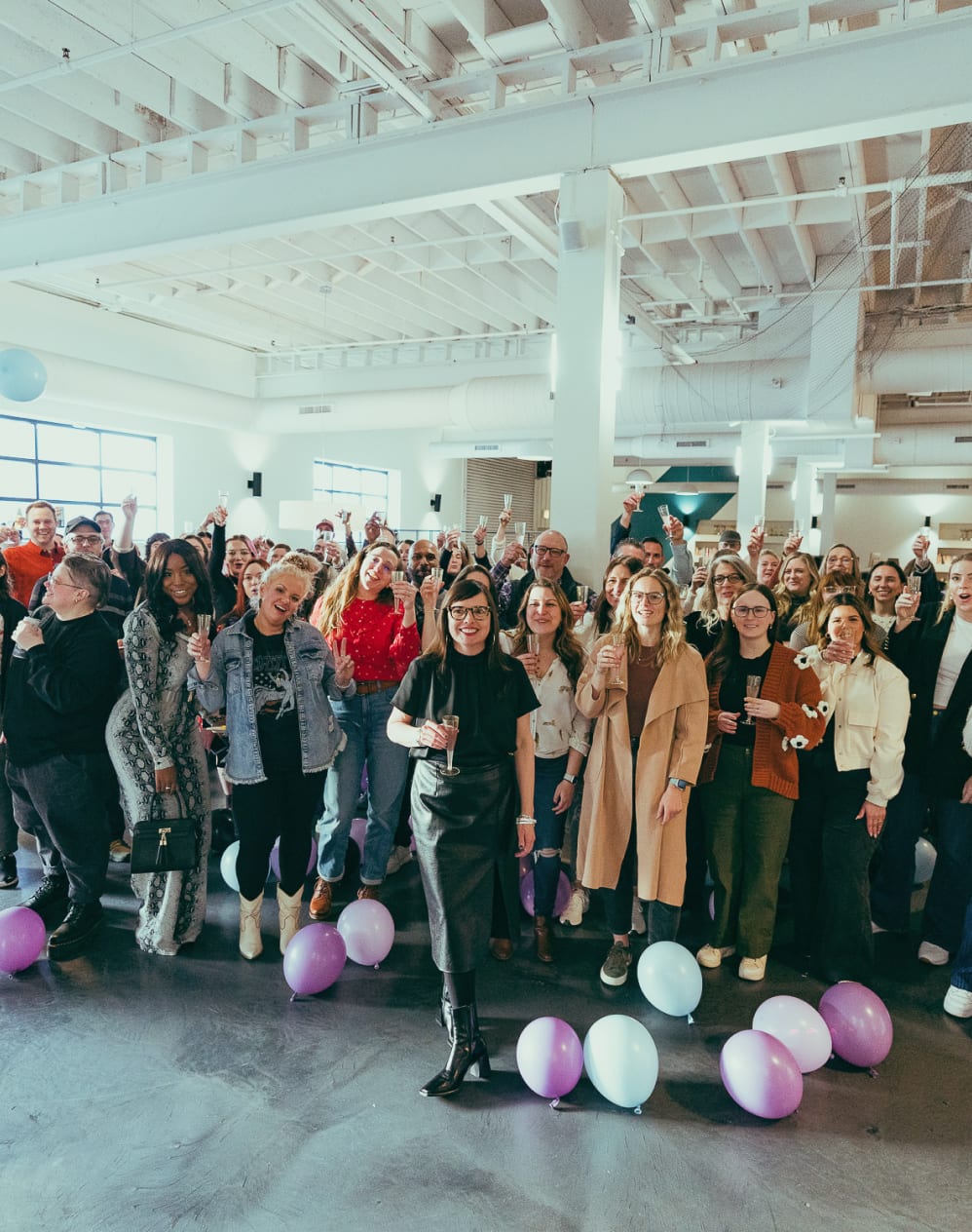 A group of people gathered in a brightly lit indoor space with balloons on the floor, some individuals holding drinks and smiling at the camera.