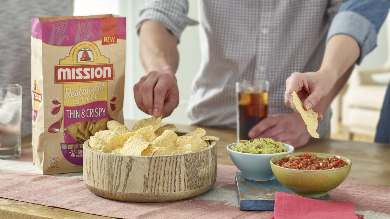 Hands serving Mission tortilla chips from a bowl with guacamole and salsa on the side