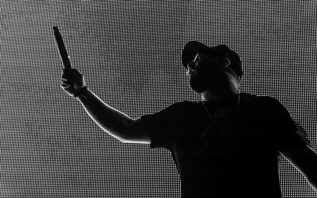 Silhouette of a man against a bright LED screen, raising his right hand and pointing upwards with a microphone in hand. He wears a cap, a T-shirt, and a necklace. The background LED screen creates a high-contrast pixelated effect around his outline.