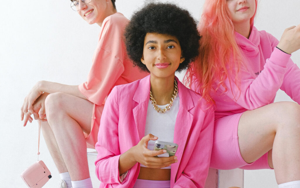 Three young women in varying shades of pink attire posing casually. The central figure, holding a smartphone, wears a pink blazer and has a curly afro, while the other two, one with glasses and the other with long pink hair, appear slightly in the background, all against a plain white backdrop.