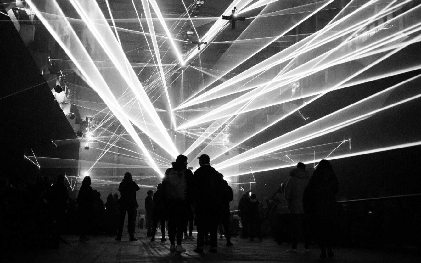 Black and white photo of people gathered in a large room, silhouetted against a dramatic display of crisscrossing laser beams shooting through the darkness, creating a dynamic and futuristic atmosphere.