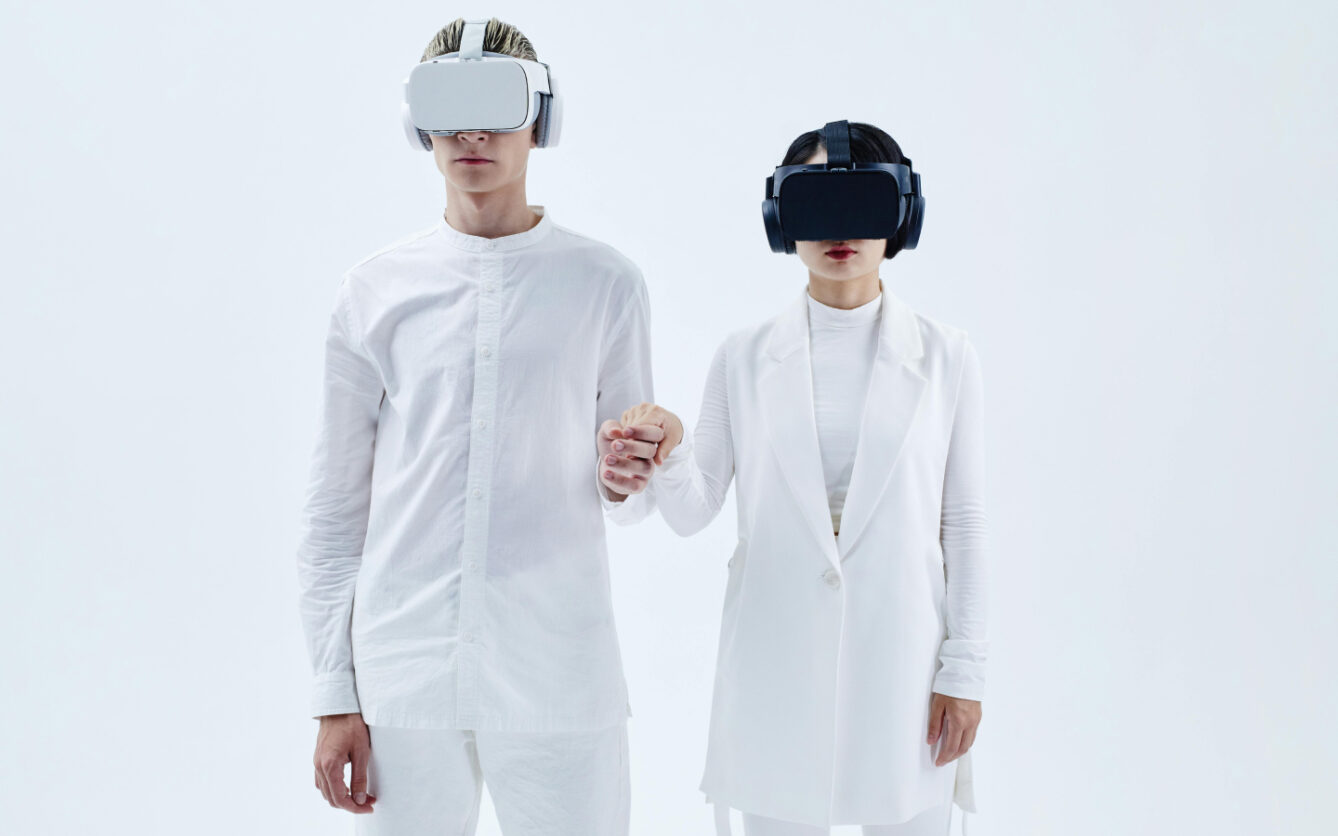 Two people wearing VR headsets and holding hands, dressed in white outfits, standing against a plain white background.