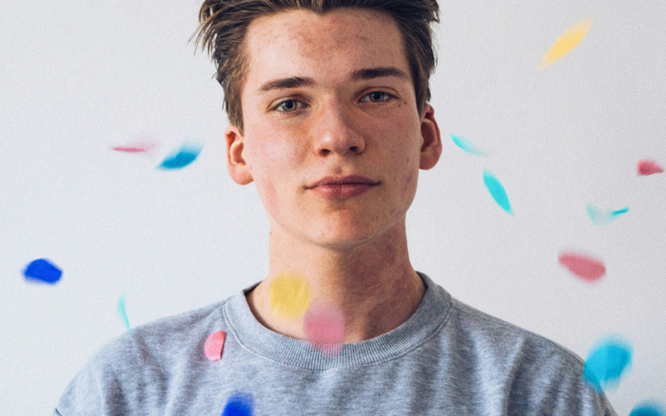Young person with short hair wearing a gray shirt, with colorful confetti floating around, against a plain white background.