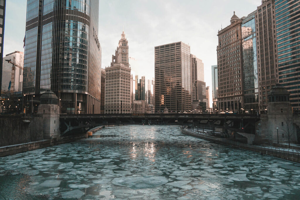 Chicago's winter skyline with buildings in the background, a partially frozen river running under a bridge.