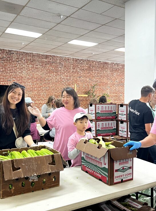 Volunteers, including a woman and child, smiling while packing corn in cardboard boxes at a community service event