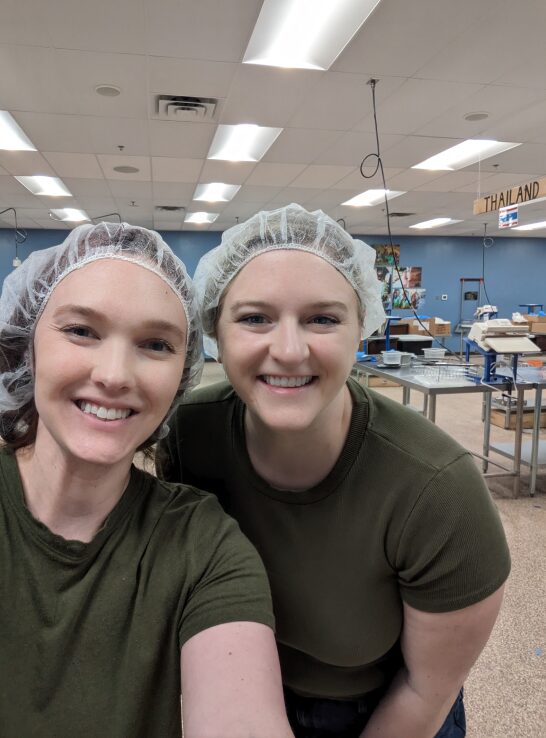 Two volunteers wearing hairnets smiling for a selfie as they are preparing food as part of a charity effort.