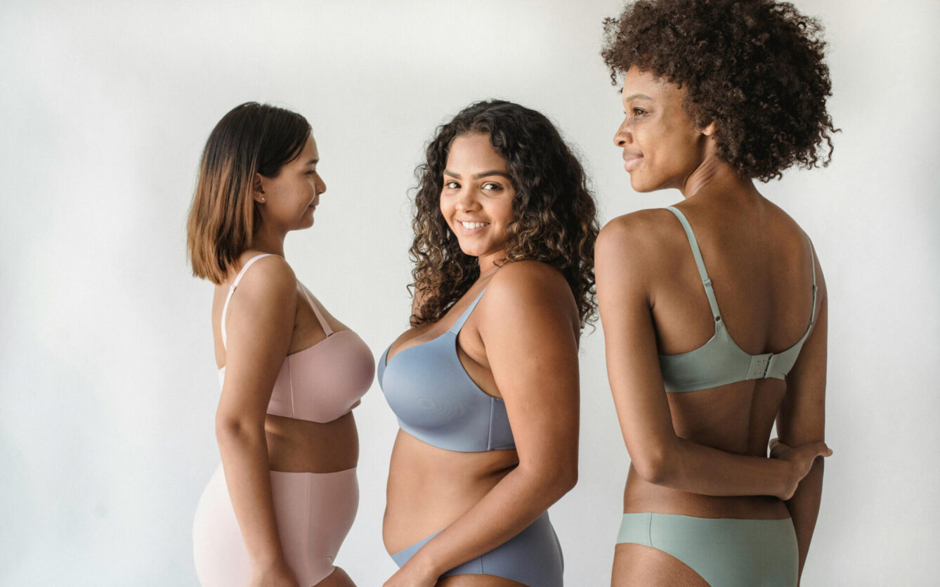 Three women of diverse body types and skin tones standing in lingerie, facing different directions and smiling. They are modeling comfortable, minimalist bras and underwear in pastel shades. The image promotes body positivity and inclusivity.