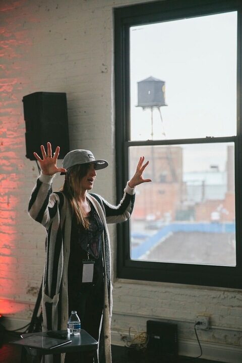 Person wearing a hat and striped sweater, standing indoors with hands raised, speaking passionately.