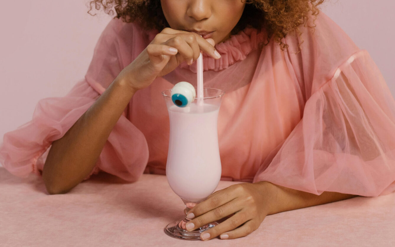 A person in a pink, sheer, ruffled top is sipping a light pink milkshake through a straw, which has a decorative eyeball garnish. The setting is playful and whimsical, matching the pastel pink theme.