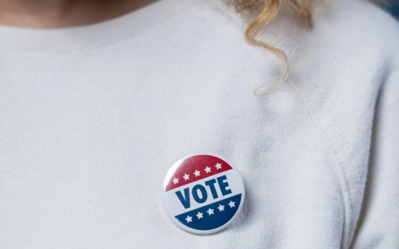 Close-up of a person wearing a white shirt with a round 'VOTE' button pinned to it. The button features red, white, and blue colors with stars, symbolizing participation in elections and civic engagement.